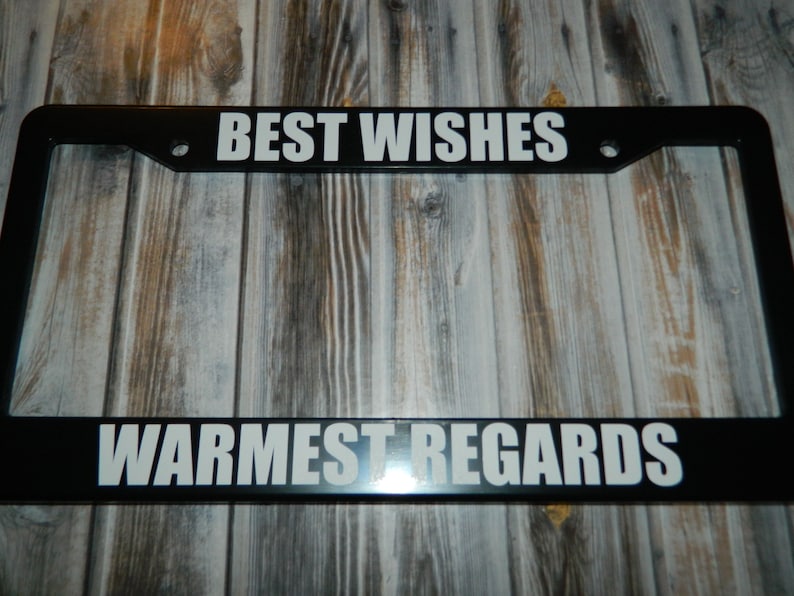 May include: Black license plate frame with white text that reads "BEST WISHES" on the top and "WARMEST REGARDS" on the bottom.