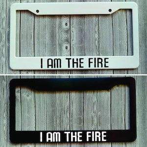 May include: Two license plate frames, one white and one black, with the text "I AM THE FIRE" printed in black on the white frame and in white on the black frame.