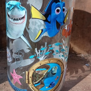 May include: Clear glass jar with a bamboo lid, featuring colourful illustrations of characters from the film "Finding Nemo." The design includes a shark, Dory, Nemo, and a sea turtle. The jar is cylindrical and sits on a wooden surface.