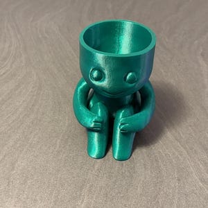 Może przedstawiać: Turkusowa doniczka wydrukowana w 3D w kształcie siedzącej postaci. Postać ma okrągłą głowę z dwoma oczami i uśmiechniętymi ustami. Jej ramiona są owinięte wokół kolan. Otwór doniczki znajduje się na szczycie głowy postaci.