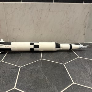 May include: A model rocket, primarily white with black and orange accents, rests on a dark gray hexagonal tile surface. The rocket features a detailed design, including engine nozzles and a pointed nose cone. The model is a replica of a Saturn V rocket.
