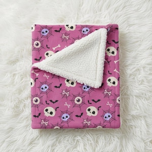 May include: A folded pink Halloween-themed blanket with a white sherpa lining. The blanket features a repeating pattern of white skulls, spiderwebs, bats, and bones. The sherpa lining is visible on the top corner of the blanket.
