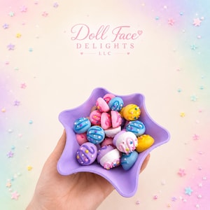 May include: A purple star-shaped bowl filled with colorful, decorated candies. The candies are various colors, including pink, blue, yellow, and purple, with white frosting and sprinkles. The image includes the text "Doll Face Delights LLC".