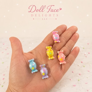 May include: Five small, candy-shaped charms in pastel colors including pink, yellow, blue, and purple. Each charm features a glossy finish with contrasting polka dot patterns. The charms are arranged in a cluster, showcasing their rounded, wrapped-candy design.