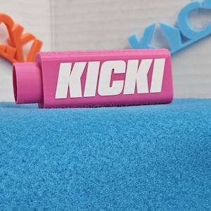 May include: A pink rectangular object with the word "KICKI" in white block letters. The object is on a blue surface. In the background, there are orange and blue lettered shapes.