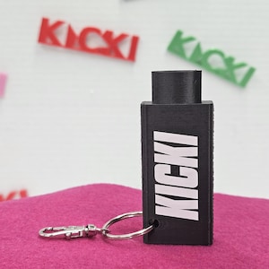Op de afbeelding: Zwarte rechthoekige sleutelhanger met het woord "KICKI" in witte blokletters. De sleutelhanger heeft een zilverkleurige sluiting en ring. De achtergrond bevat het woord "KICKI" in rood, groen en roze.