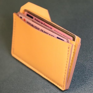May include: A yellow leather card holder with visible stitching. The holder contains several cards, including a French driver's license. The words "REPUBLIQUE FRANCAISE" are visible on one of the cards.