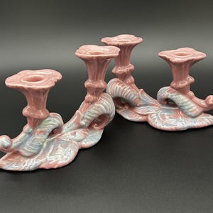 May include: Set of two ceramic candle holders. Each holder features a pink, floral-shaped cup and a base with a swirling, leaf-like design in pink, blue, and white. The candle holders are on a black surface.