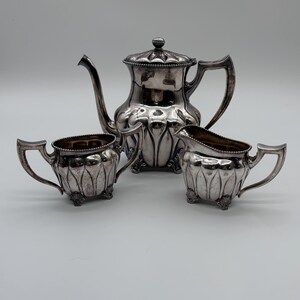 May include: A silver-plated tea set consisting of a teapot with a lid, a sugar bowl with two handles, and a creamer pitcher. Each piece features a scalloped, petal-like body design, beaded rim detailing, and small decorative feet at the base.
