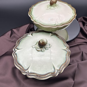 May include: A two-piece ceramic serving dish with a lid. The dish and lid are light green with a scalloped edge and a brown trim. The lid features a brown acorn-shaped handle. The dish is on a reflective surface.