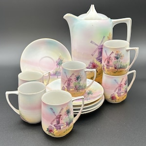 May include: A porcelain tea set featuring a tall teapot and four mugs with matching saucers. Each piece displays a hand-painted scene of a windmill in a landscape with soft pastel colors, including pink, blue, and yellow, finished with gold trim.