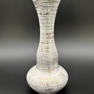 Op de afbeelding: Een hoge, witte keramische vaas met een gestructureerd, horizontaal streepontwerp. De vaas heeft een bolvormige basis, een slanke hals en een uitlopende bovenkant. Gouden accenten zijn zichtbaar op de rand en de basis. Een decoratief woondecoratie-item.