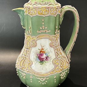 May include: A vintage green porcelain coffee pot with a lid, handle, and spout. The pot features ornate yellow and red trim, and a central panel with a floral bouquet design. The pot is a decorative item.