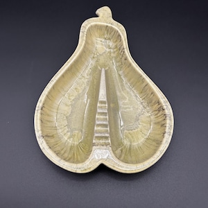 May include: A pear-shaped, two-sectioned ceramic dish in shades of green and tan. The dish has a raised center divider and a small, curved handle at the top. The surface has a marbled appearance.