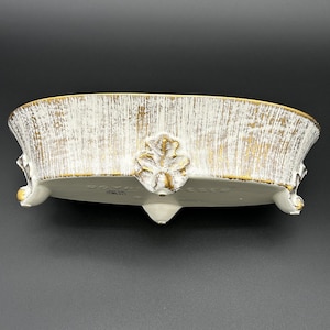 Könnte beinhalten: Eine dekorative Schale aus weißer und goldener Keramik mit strukturierter Oberfläche. Die ovale Schale hat goldene Verzierungen und Blattmotive. Die Schale wird von drei kleinen Füßen getragen und hat ein Blattdesign auf der Vorderseite.