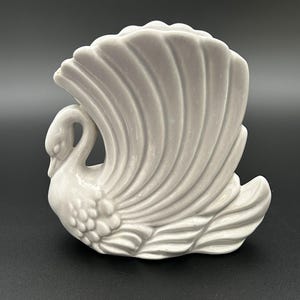 Op de afbeelding: Een witte keramische zwaanfiguur met een gedetailleerd veermotief. De zwaan heeft een gebogen nek en staat met gespreide vleugels. De figuur is een decoratief item.