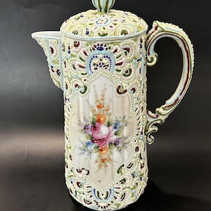 以下が含まれることがあります： 蓋、取っ手、注ぎ口が付いた、装飾的なアンティークのセラミックコーヒーポット。 ポットはクリーム色で、赤、青、緑、黄色のカラフルな花柄とスクロールワークのデザインが施されています。 中央のパネルには花のブーケが描かれています。