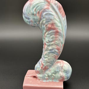 May include: A ceramic sculpture with a curved, abstract form. The sculpture features a blend of colours, including pink, blue, and red, with a marbled effect. It rests on a rectangular pink base, creating a striking contrast. The piece has a smooth, glossy finish.