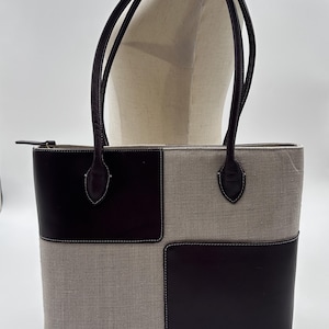 May include: A rectangular tote bag featuring a color-blocked design with alternating panels of dark brown leather and light beige textured fabric. The bag includes two dark brown leather shoulder straps and a small gold-toned logo on the bottom right.