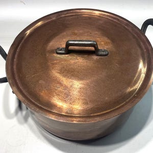 May include: A copper cooking pot with a lid. The pot features two black, curved handles and a small handle on the lid. The copper has a warm, reddish-brown hue, and the pot shows signs of use.