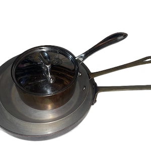 May include: A set of three stacked metal pans with long handles. The top pan is a small copper-toned saucepan with a polished stainless steel lid. Beneath it are two larger, flat, circular metal pans with long, slender, metallic handles.