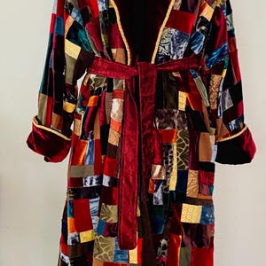 Pode incluir: Um roupão de patchwork em uma variedade de cores, incluindo vermelho, bordô, dourado e azul. O roupão tem gola e punhos de veludo bordô, com acabamento dourado. Um cinto bordô combinando afivela a cintura. O roupão é feito de vários tecidos de veludo.
