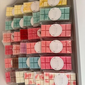 May include: A selection of colourful wax melts, including yellow, teal, pink, and white, presented in a tray. Each melt is individually wrapped in clear plastic and labelled with a circular sticker reading "THE FLAME". The melts are divided into small squares.
