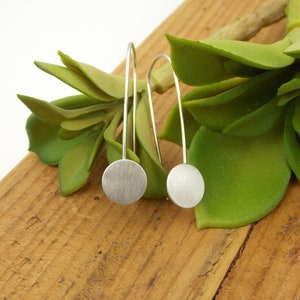 May include: A pair of silver dangle earrings with round, brushed silver discs. The earrings are photographed on a wooden surface with a green succulent plant.