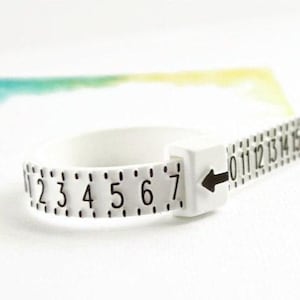 May include: A white plastic ring sizer featuring black printed numbers and tick marks along the band. The device includes a square housing with a black arrow indicator pointing toward the numerical scale to determine the circumference of a finger.