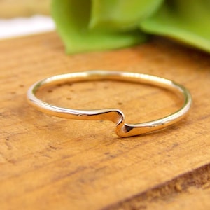 Puede incluir: Un delicado anillo de oro con un diseño de onda único. El anillo se encuentra sobre una superficie de madera, resaltando su acabado pulido. El estilo minimalista del anillo lo convierte en un accesorio versátil.