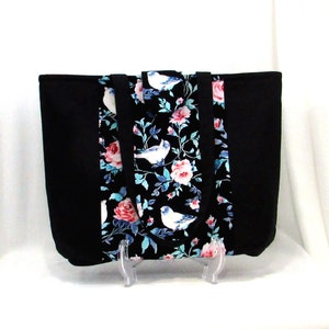 May include: Black tote bag with a floral print featuring pink roses and white birds on a teal background. The bag has black fabric on the sides and a black handle.