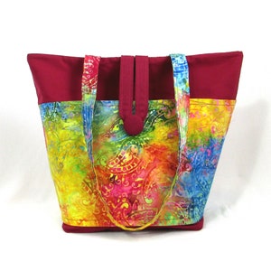 May include: A large tote bag with a red top and a colorful batik fabric bottom. The bag has two long straps and a flap closure.