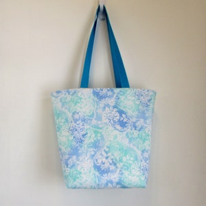 May include: A blue and white floral tote bag with turquoise handles. The bag is hanging from a hook on a white wall.