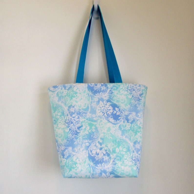 May include: A blue and white floral tote bag with turquoise handles. The bag is hanging from a hook on a white wall.