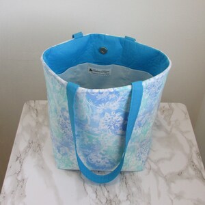 May include: A blue and white floral tote bag with turquoise handles. The bag is made of fabric and has a white lining. The bag has a small label that says "Baltimore Designs Handmade purses & bags".