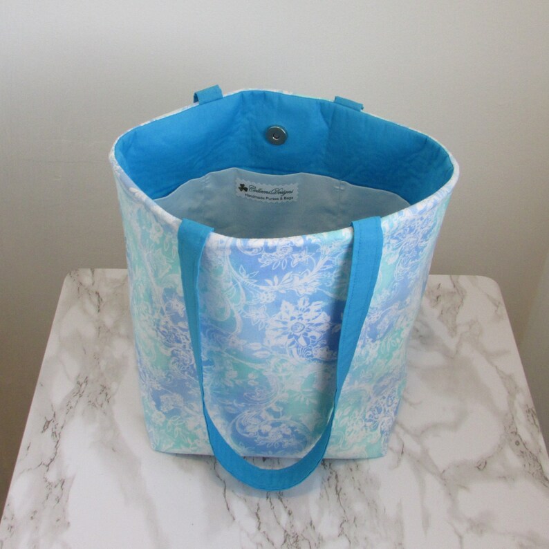 May include: A blue and white floral tote bag with turquoise handles. The bag is made of fabric and has a white lining. The bag has a small label that says "Baltimore Designs Handmade purses & bags".