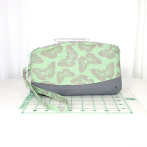 May include: A rectangular clutch with a light green butterfly pattern on the top half and a solid gray bottom. The clutch has a matching wrist strap and a zipper closure. The background is white.