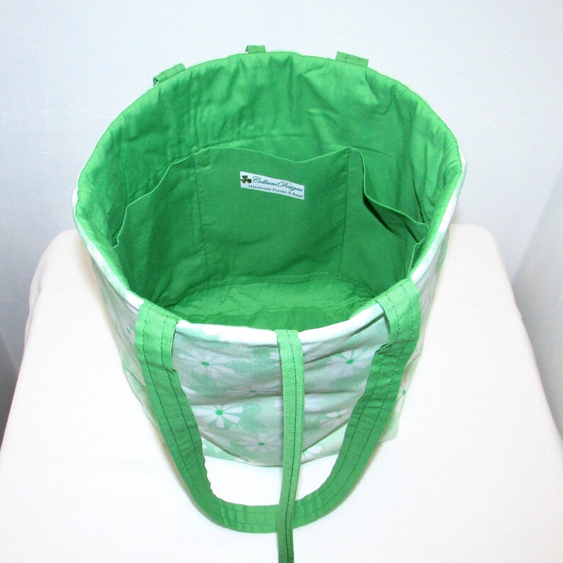 May include: Green tote bag with white floral print and green straps. The bag has a green lining with two pockets. The bag has a label that reads "Ballroom Designs Handmade Quilts & Bags".