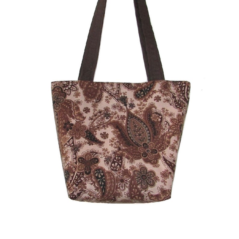 Pode incluir: Uma sacola com um padr&atilde;o paisley em tons de marrom e bege. A sacola tem formato retangular com uma abertura larga e duas al&ccedil;as marrons escuras. O tecido apresenta um design floral e em forma de gota repetitivo.