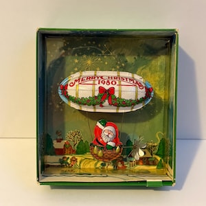 Könnte beinhalten: Ein weihnachtlicher Vintage-Schmuck aus dem Jahr 1980, bestehend aus einem weiß-blauen Luftschiff mit der Aufschrift MERRY CHRISTMAS 1980, verziert mit Stechpalmen und einer roten Schleife. Ein Weihnachtsmann sitzt in einem braunen Korb.