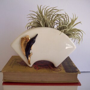 May include: White ceramic planter shaped like a fan with a black and gold marlin design. The planter is filled with a green air plant.