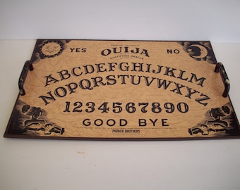May include: Ouija board with a light brown wood grain finish. The board features the alphabet, numbers, and the words "Yes," "No," and "Goodbye." Decorative elements include a sun, moon, and stylized figures. The board has two handles.