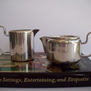 May include: A stainless steel tea set, including a small teapot with a lid and a creamer. The teapot and creamer have curved handles and spouts. The set is sitting on a book with the title "Table Settings, Entertaining, and Etiquette".