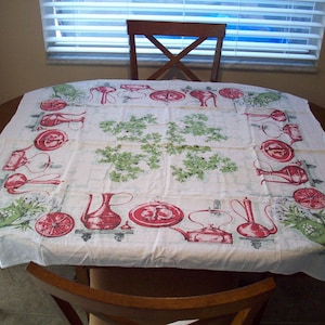 May include: A white tablecloth with a repeating pattern of red kitchen tools and green floral accents. The tablecloth is draped over a round wooden table, creating a vintage aesthetic. The design includes teapots, pitchers, and other kitchen items.