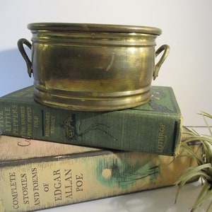 May include: A small, round brass planter with two handles. The planter is sitting on top of two books. The top book is green and has the text "LOTHROP" on the spine. The bottom book is cream colored and has the text "COMPLETE STORIES AND POEMS OF EDGAR ALLAN POE" on the spine.