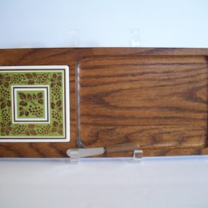 May include: A rectangular wooden serving board with a recessed area and a square ceramic tile inlay featuring a green and brown grape vine pattern. A small metal spreader with a wooden handle is positioned horizontally along the bottom edge.