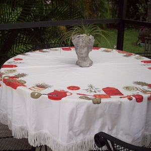 May include: A white tablecloth with a red and green border featuring a pattern of kitchen utensils. A potted plant in a bust-shaped planter sits in the center of the table.