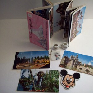 May include: A collection of vintage Disneyland postcards featuring images of the park, including Sleeping Beauty Castle, the Matterhorn, and the Pirates of the Caribbean ride. There are also two small white teacups and a Mickey Mouse pin.