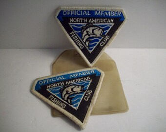 May include: Two triangular embroidered patches featuring a black background, a blue border, and white text reading OFFICIAL MEMBER NORTH AMERICAN FISHING CLUB. Each patch displays a white graphic of a fish jumping over blue waves.