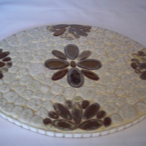 May include: A round, white, ceramic coaster with a mosaic design. The coaster features three brown flower-shaped designs made of small, oval-shaped tiles.